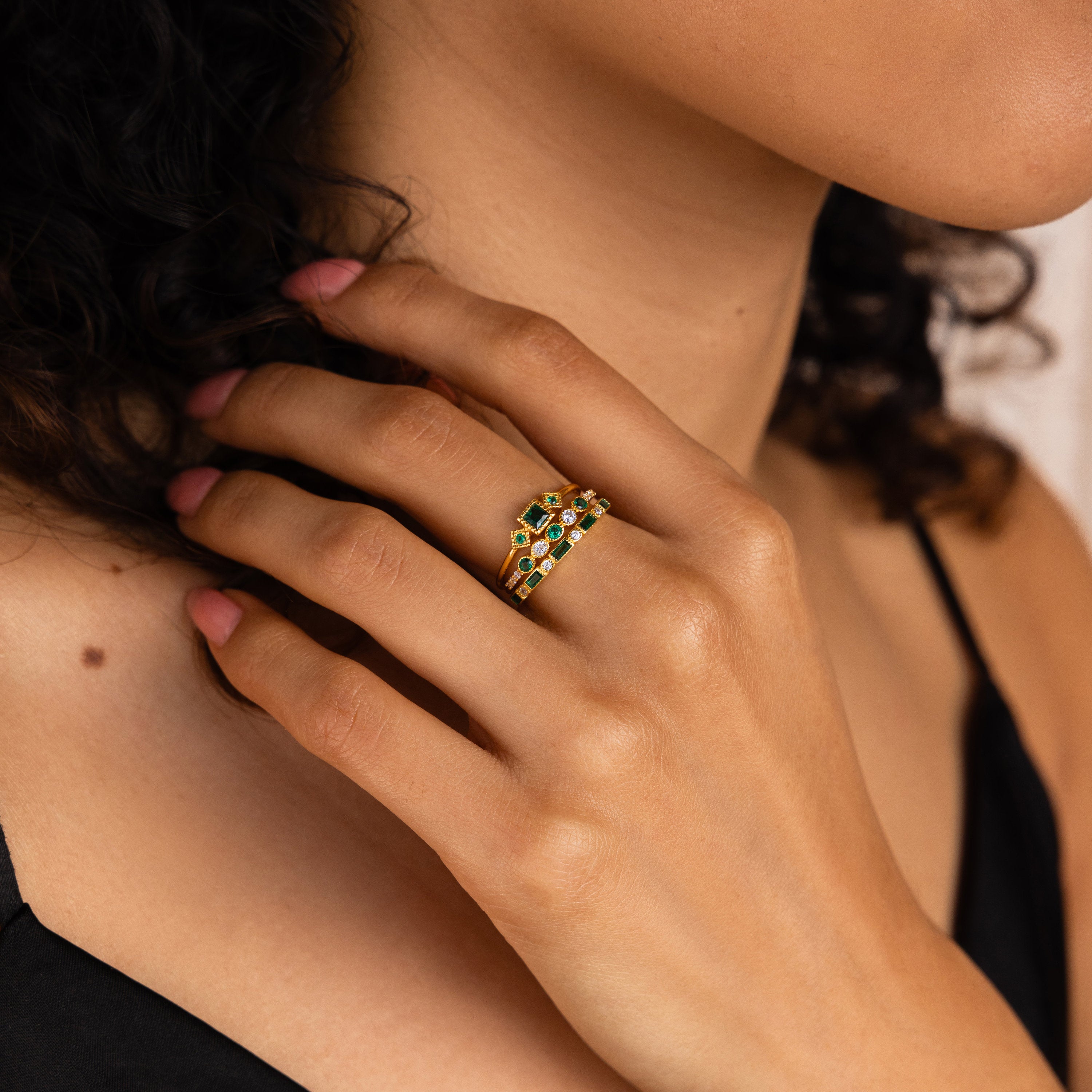 Woman with curly hair wearing the Emerald Stacking Ring Set in 18K Gold, touching her face while dressed in a black top.