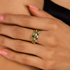 A hand with pink polished nails wears two gold rings, including the Emerald Stacking Ring Set with green and clear stones, showcasing vintage-inspired elegance against bare skin.