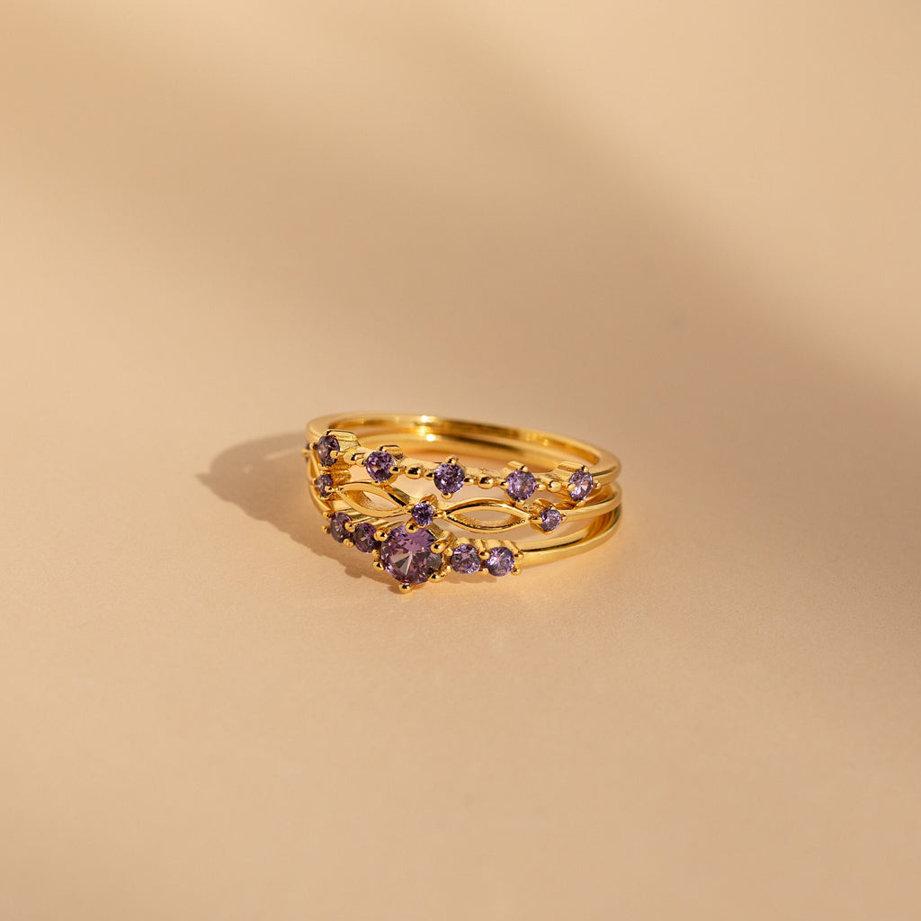 The Amethyst Stacking Ring Set features gold rings with purple gemstones in a delicate pattern on a beige background—ideal for stacking or wearing as a striking statement piece.