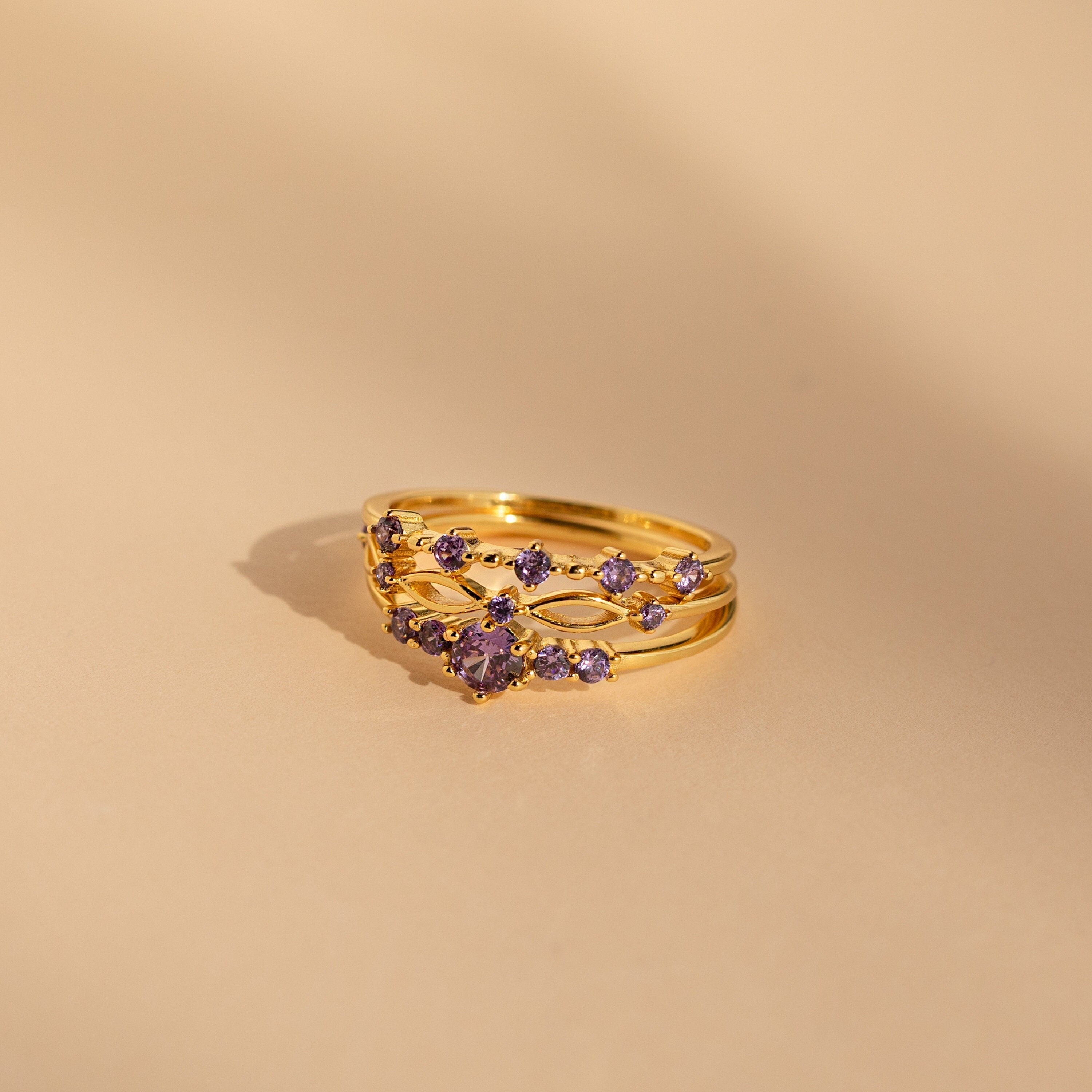 The Amethyst Stacking Ring Set features gold rings with purple gemstones in a delicate pattern on a beige background—ideal for stacking or wearing as a striking statement piece.