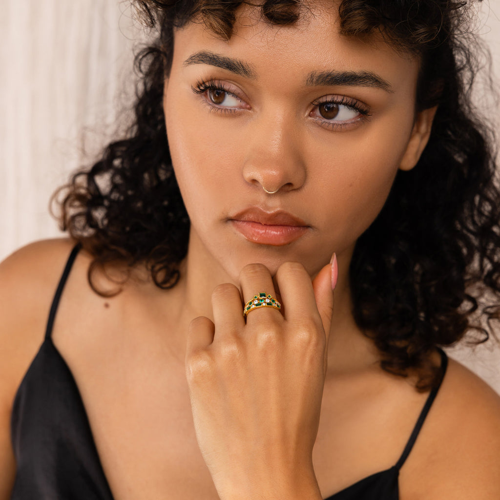 A woman with curly hair in a black top wears the Emerald Stacking Ring Set, gazing thoughtfully to the side—her look is elegantly finished with this vintage-inspired art deco jewelry.