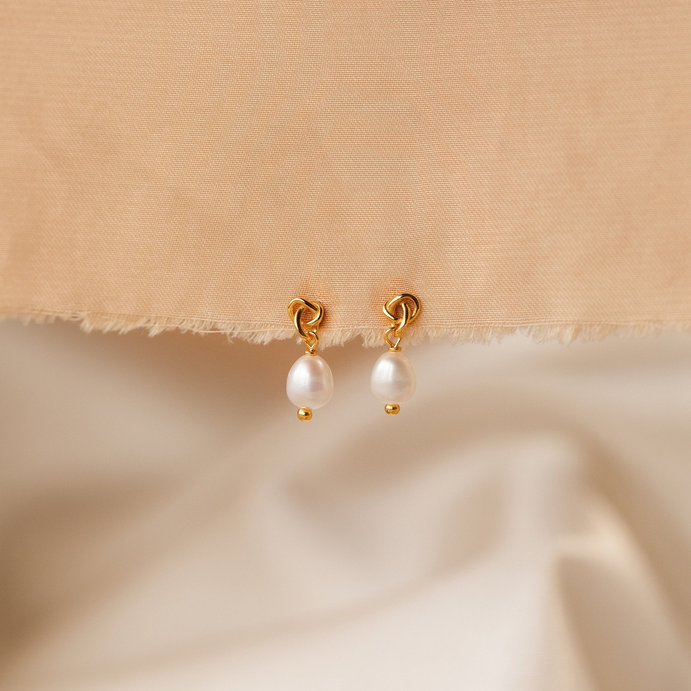 The Dainty Knot Pearl Earrings in 18K Gold exude timeless sophistication, beautifully showcased on a beige textured fabric background.