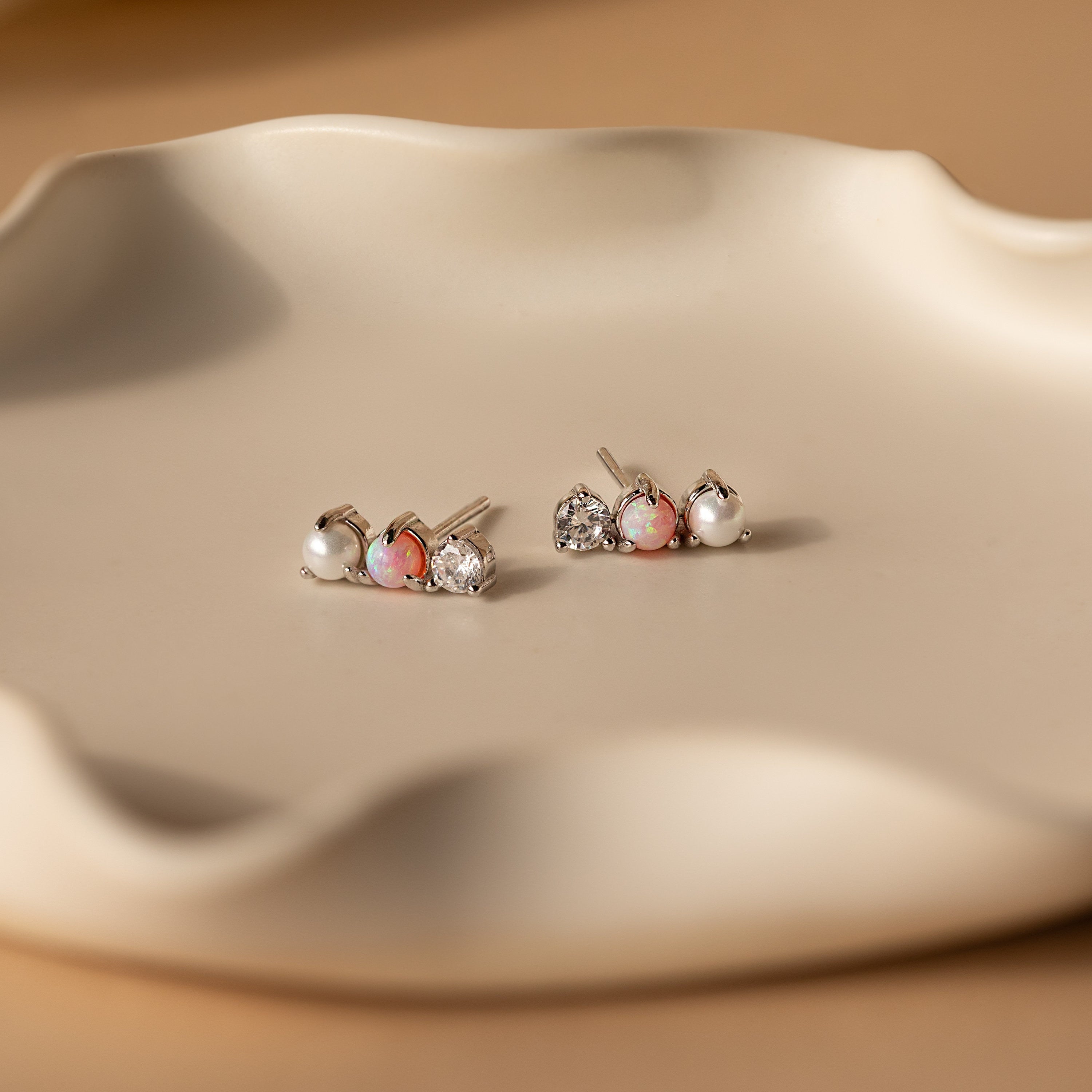 Three pairs of Pearl, Opal & Diamond Studs are displayed on a wavy beige ceramic dish.