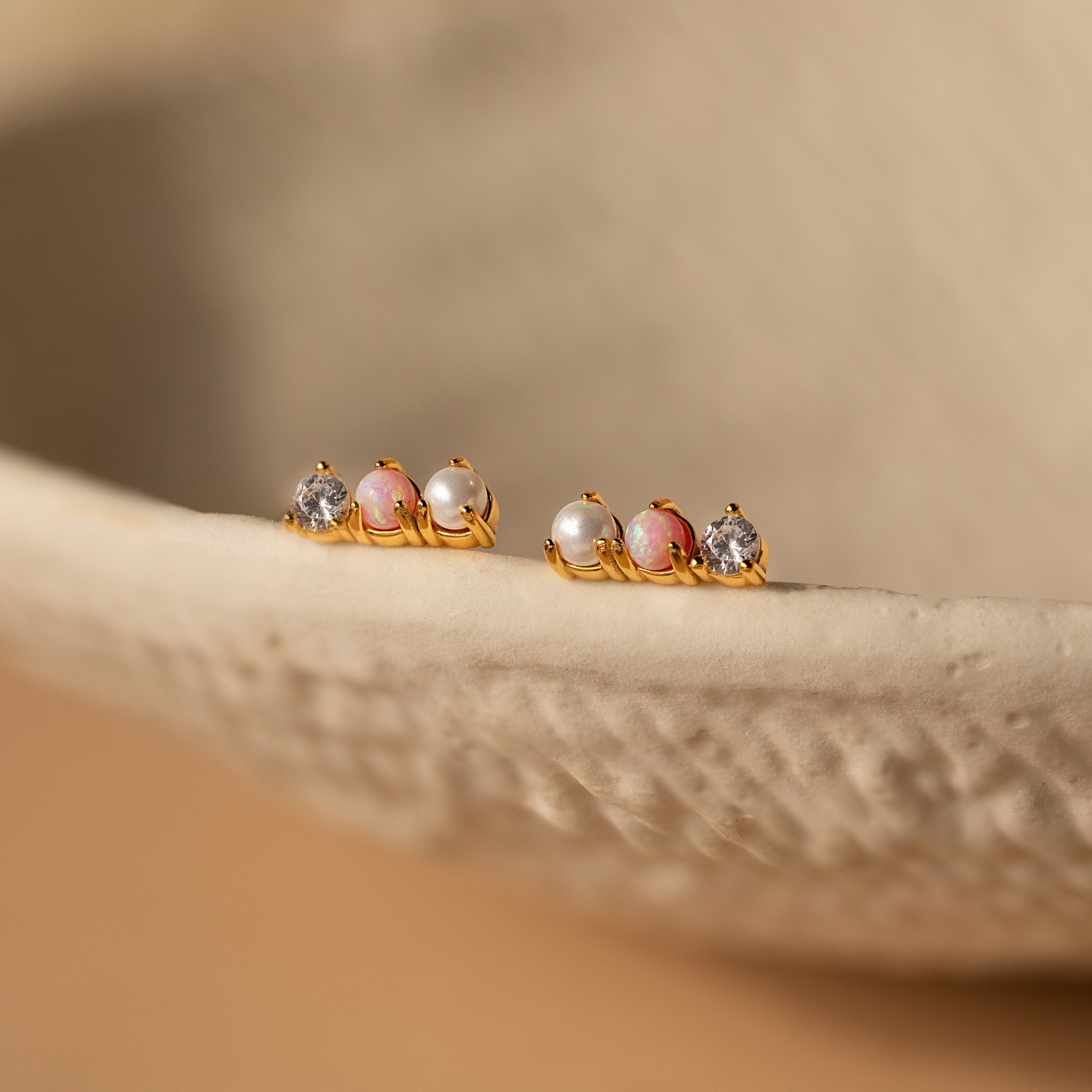 Two pairs of Pearl, Opal & Diamond Studs with pink stones and clear gems rest on a textured beige surface, offering a delicate alternative to classic diamond studs.