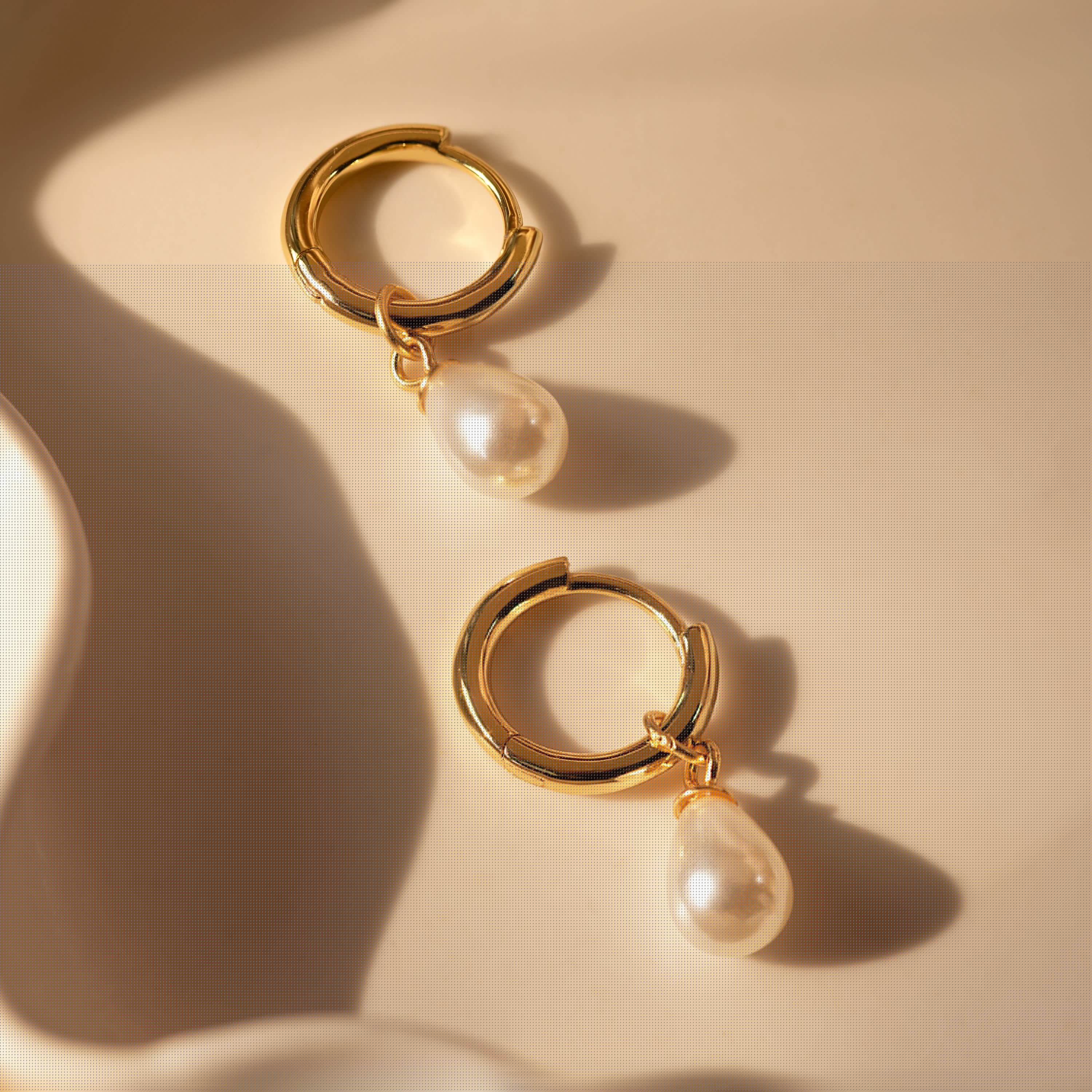 Dainty Pearl Drop Huggies: Elegant gold hoop earrings featuring teardrop pearls, shown on a beige surface with soft shadows—ideal for adding a refined touch to any occasion.