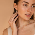 A woman wearing Pearl Threader Earrings in minimalistic gold touches her earring and gazes to the side against a beige background.