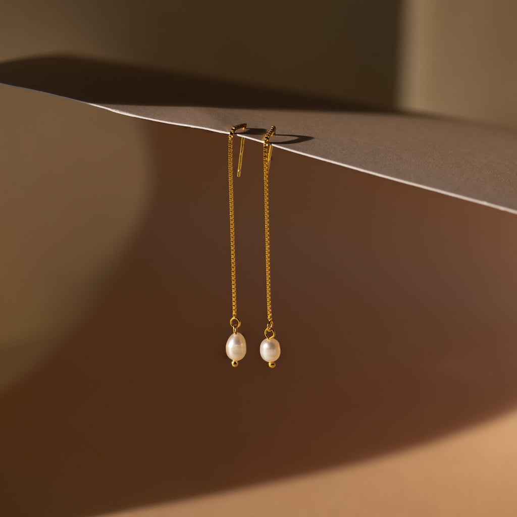 The Pearl Threader Earrings feature a minimalistic gold design with pearls delicately suspended from a chain, creating elegant dangling charm earrings.
