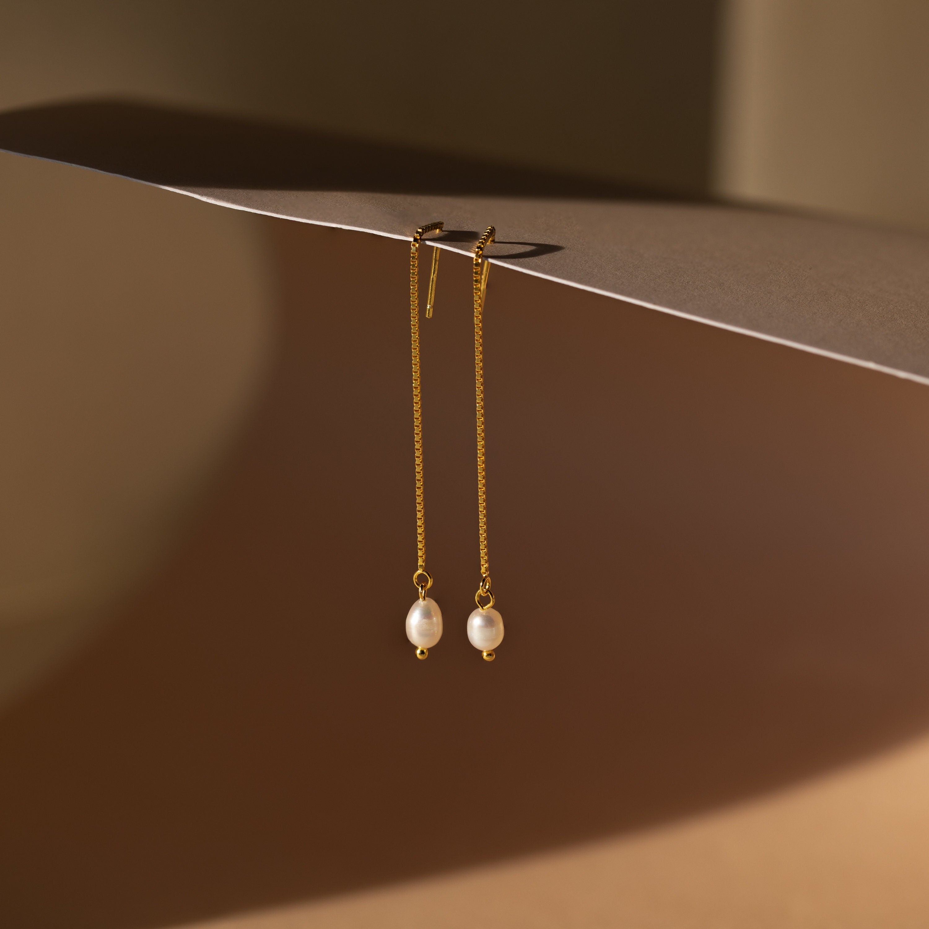 The Pearl Threader Earrings feature a minimalistic gold design with pearls delicately suspended from a chain, creating elegant dangling charm earrings.