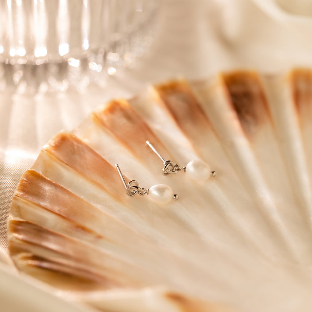 Dainty Knot Pearl Earrings in Sterling Silver displayed on a brown and cream seashell with a glass in the background, highlighting their classic charm and timeless sophistication.