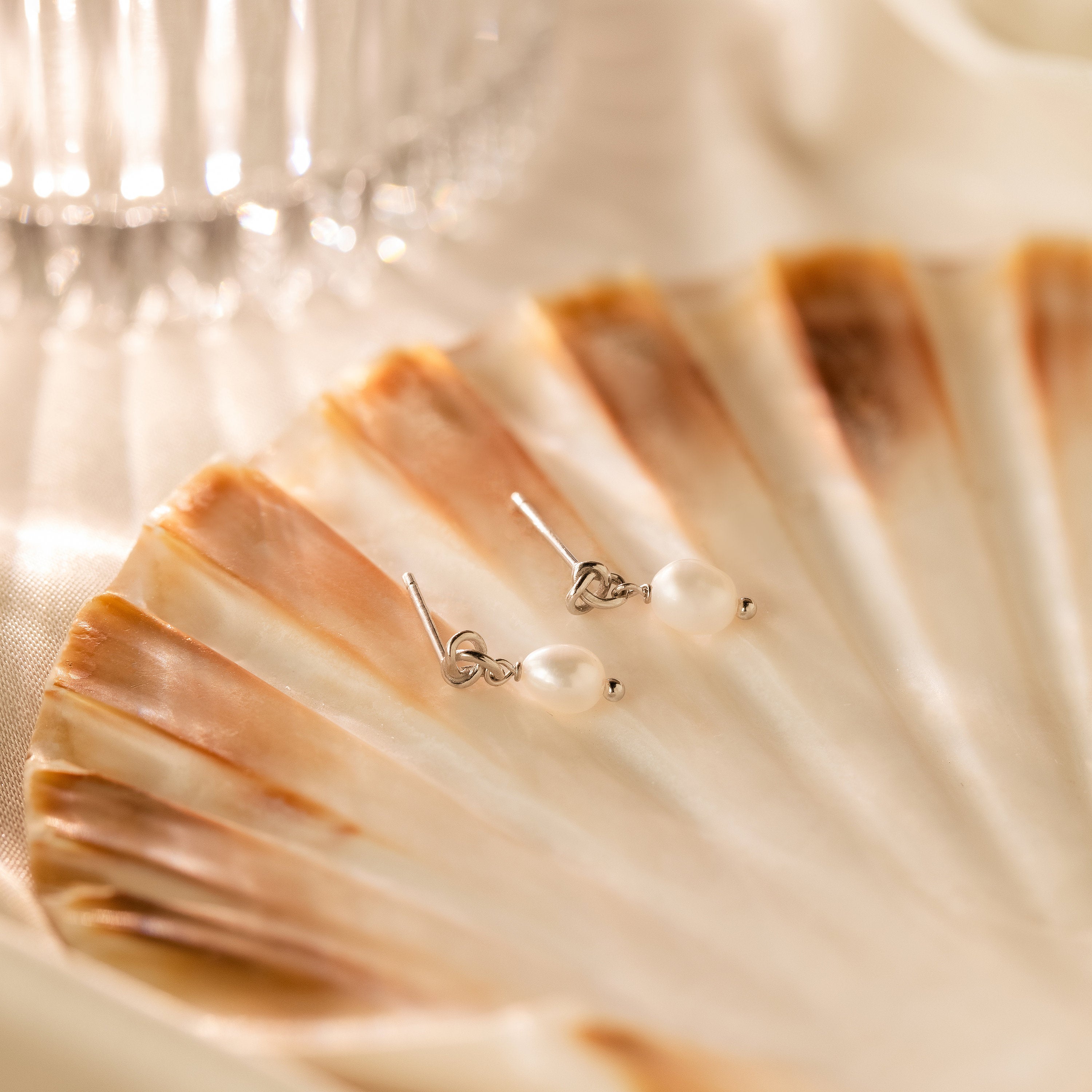 Dainty Knot Pearl Earrings in Sterling Silver displayed on a brown and cream seashell with a glass in the background, highlighting their classic charm and timeless sophistication.