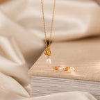 The Gold necklace with a pearl pendant and Dainty Knot Pearl Earrings, displayed on a closed beige book, evoke timeless sophistication.