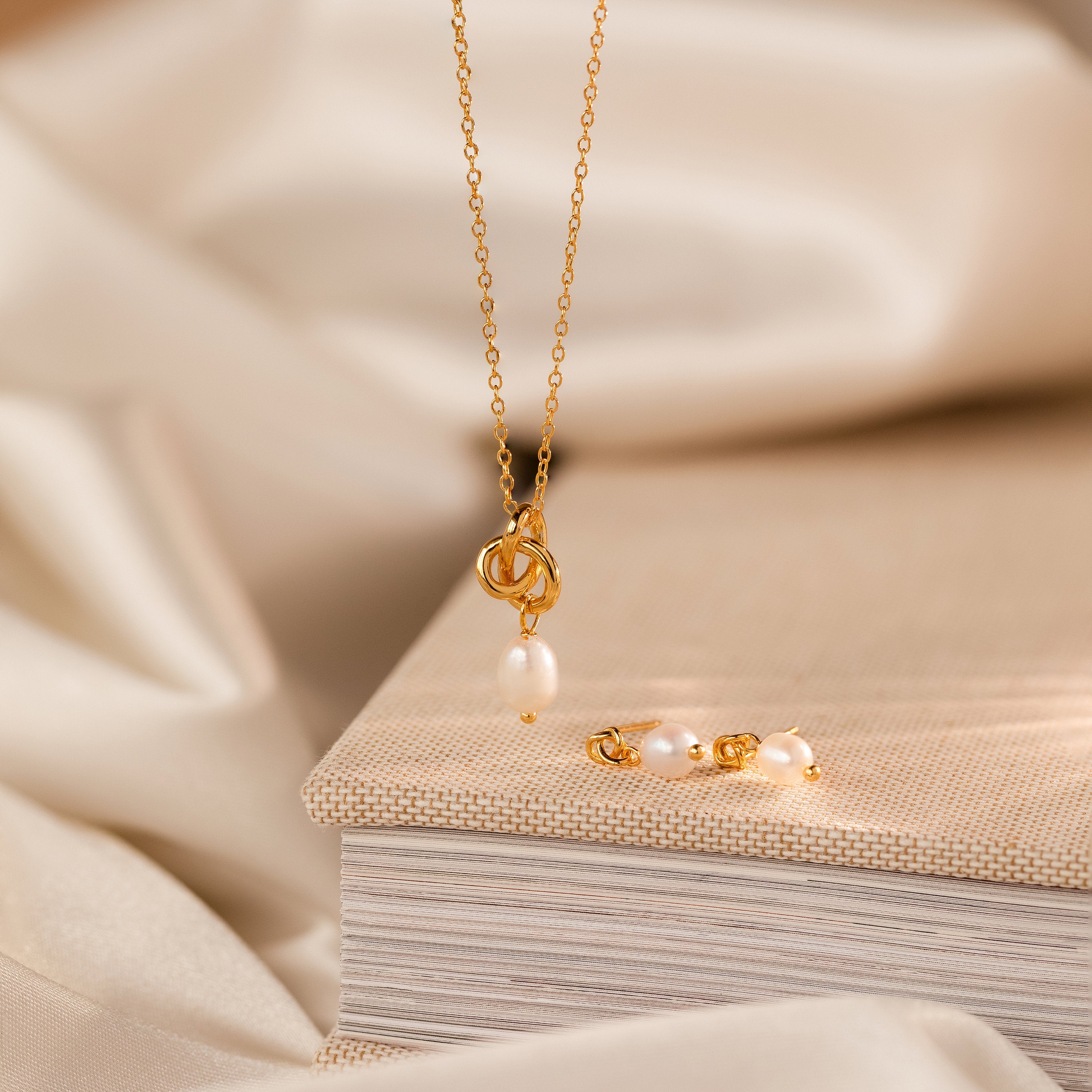The Gold necklace with a pearl pendant and Dainty Knot Pearl Earrings, displayed on a closed beige book, evoke timeless sophistication.