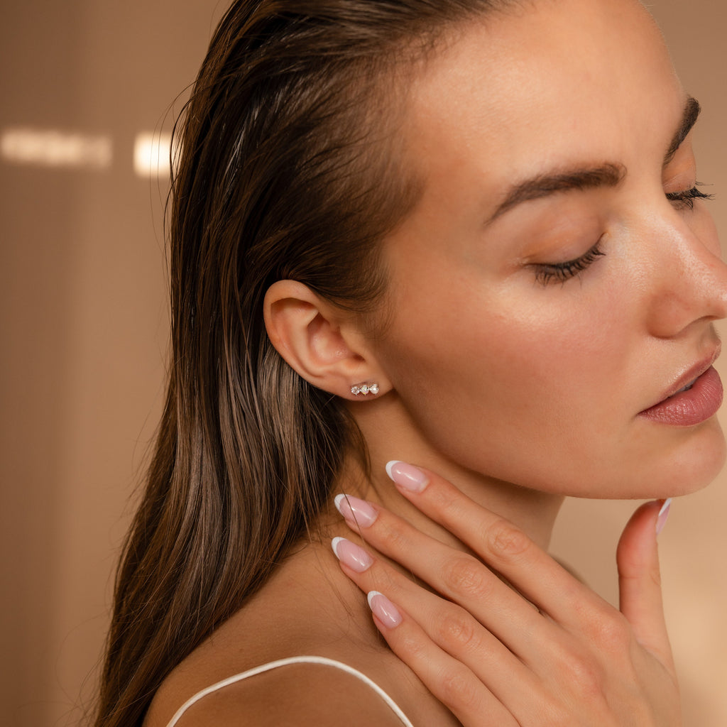 A woman with closed eyes touches her neck, highlighting Pearl, Opal & Diamond Studs and a nude manicure against a beige background.