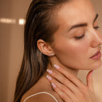 A woman with closed eyes touches her neck, highlighting Pearl, Opal & Diamond Studs and a nude manicure against a beige background.