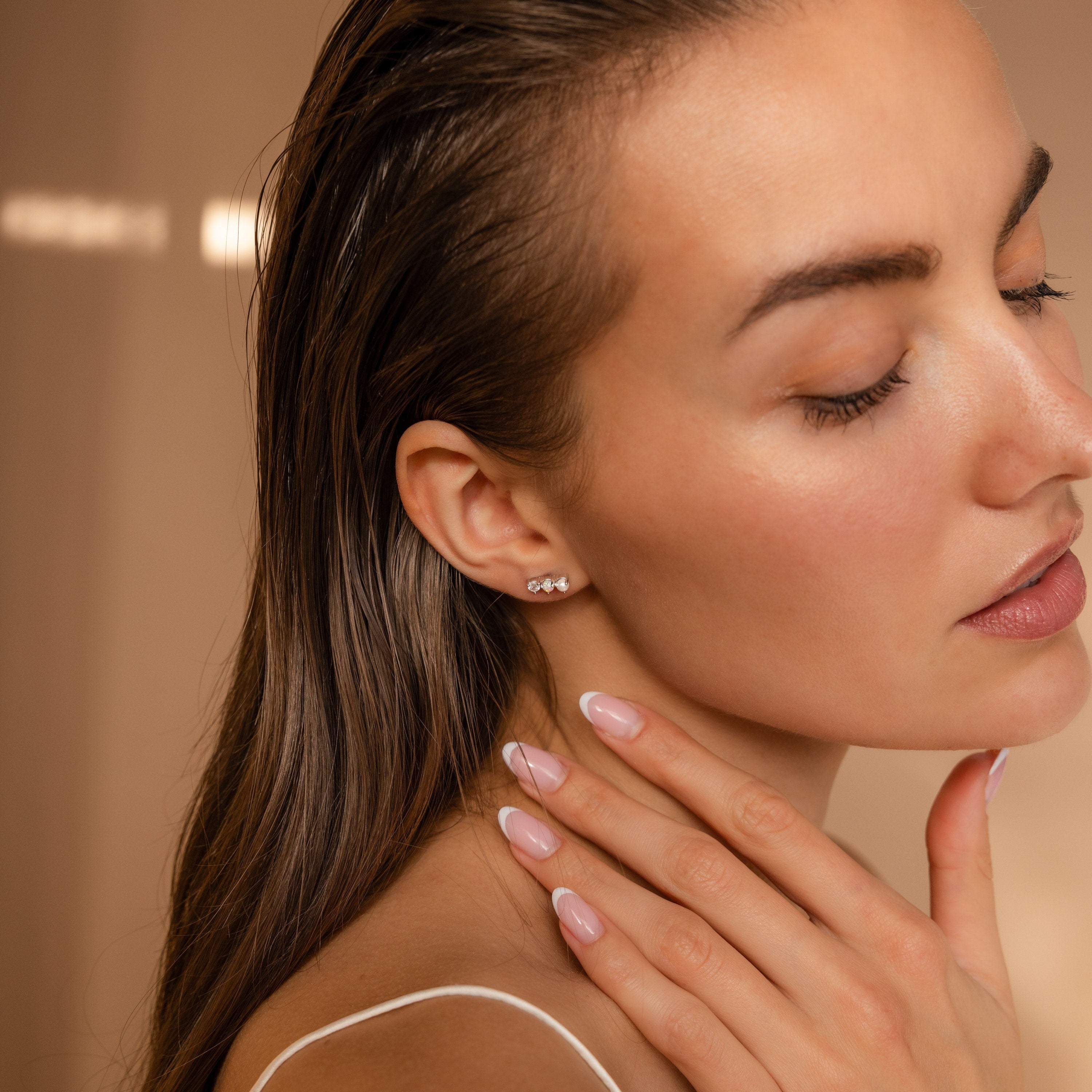 A woman with closed eyes touches her neck, highlighting Pearl, Opal & Diamond Studs and a nude manicure against a beige background.