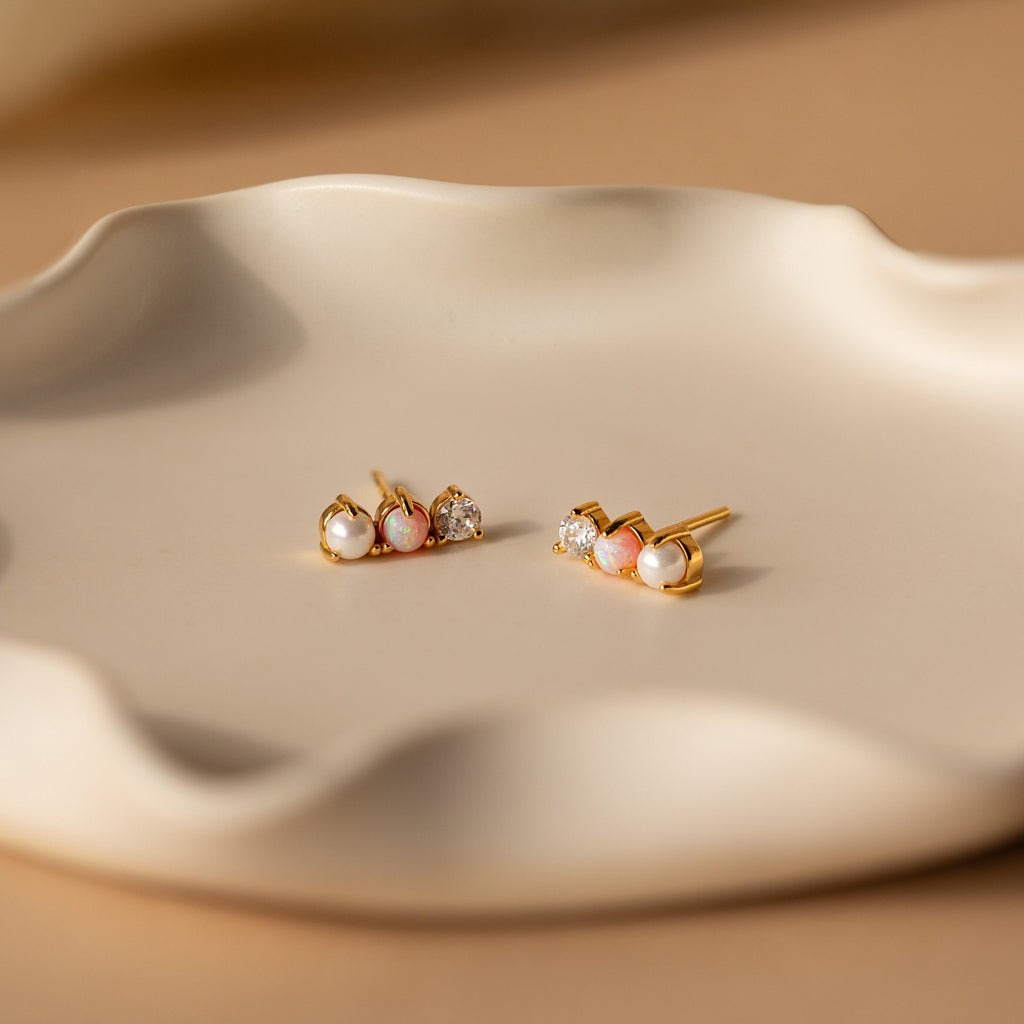 Pearl, Opal & Diamond Studs rest on a wavy, cream-colored ceramic dish in soft lighting.