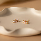Pearl, Opal & Diamond Studs rest on a wavy, cream-colored ceramic dish in soft lighting.