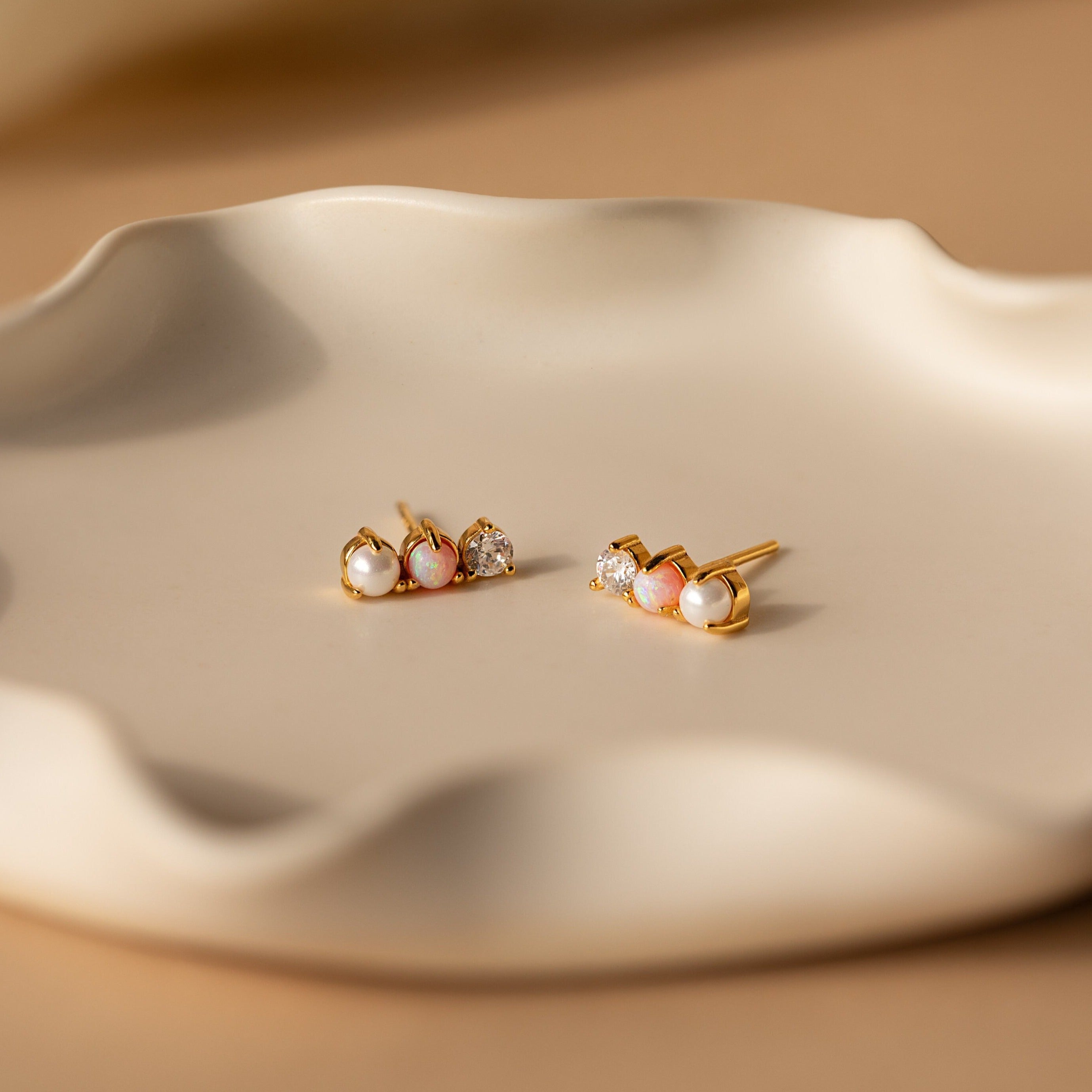 Pearl, Opal & Diamond Studs rest on a wavy, cream-colored ceramic dish in soft lighting.