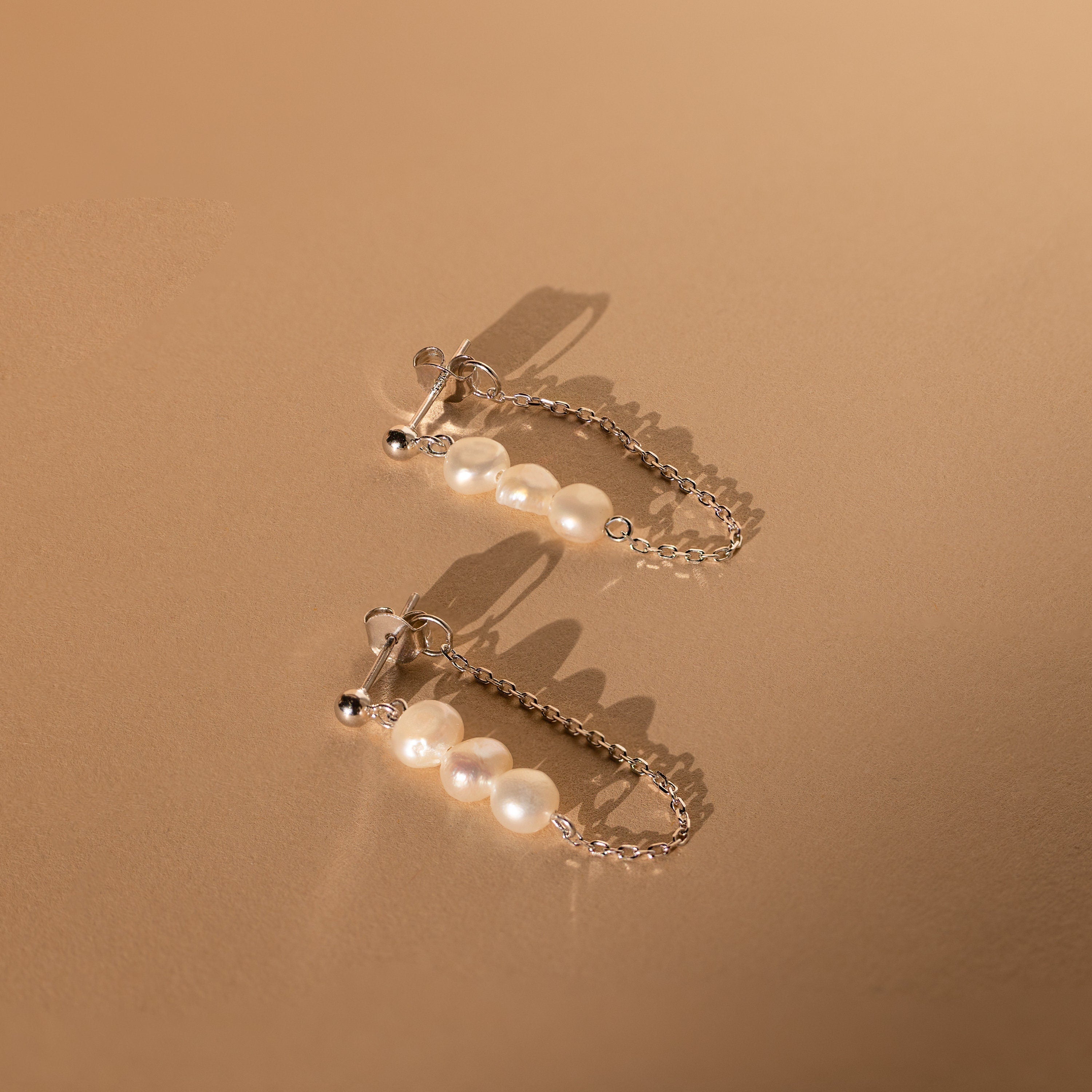 Two elegant Triple Pearl Drop Earrings with pearls and silver chains are displayed on a neutral beige background, perfect for fans of refined jewelry.