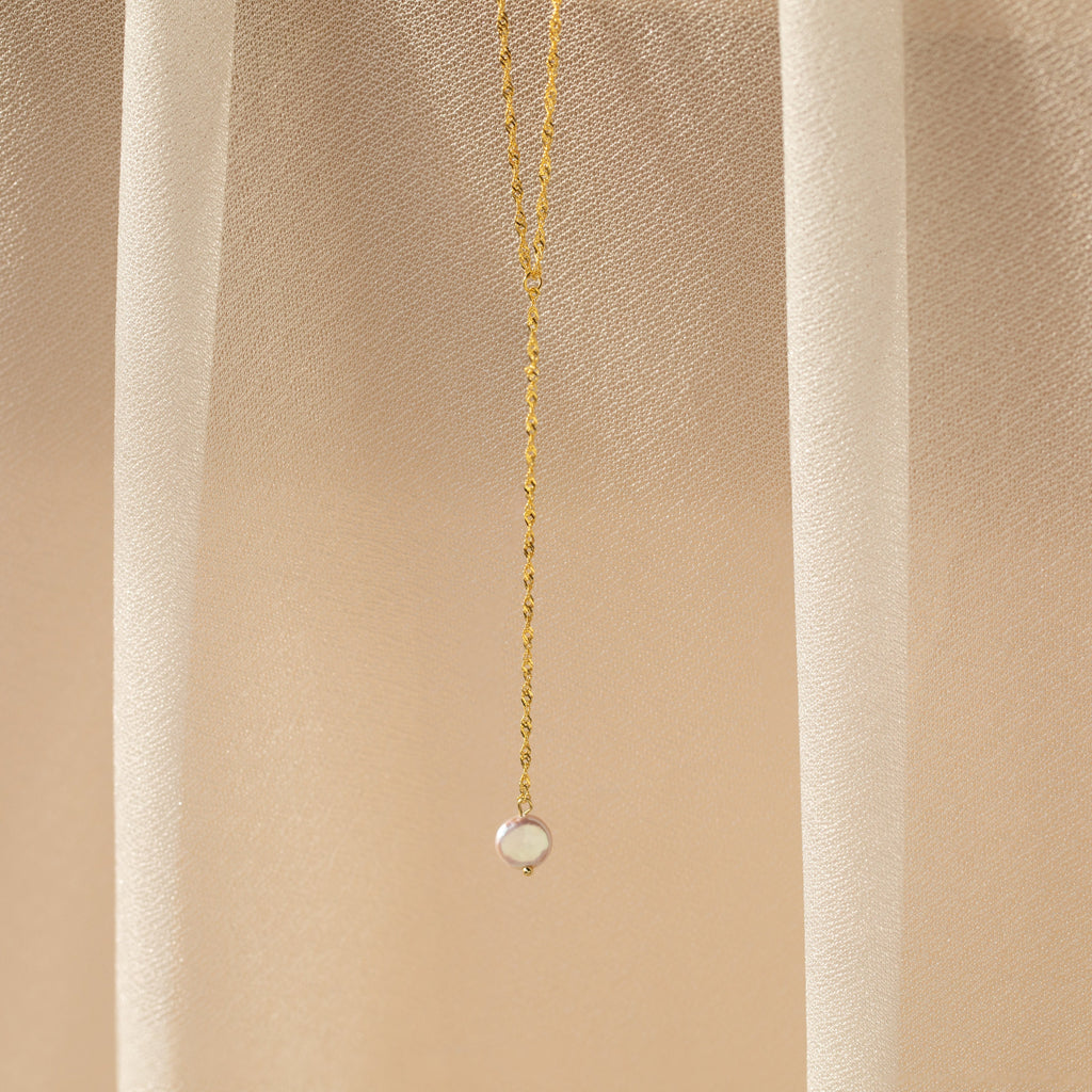 A delicate chain featuring a small round pendant is displayed on a beige, sheer fabric—perfect for fans of timeless elegance like the Pearl Lariat Y Necklace.