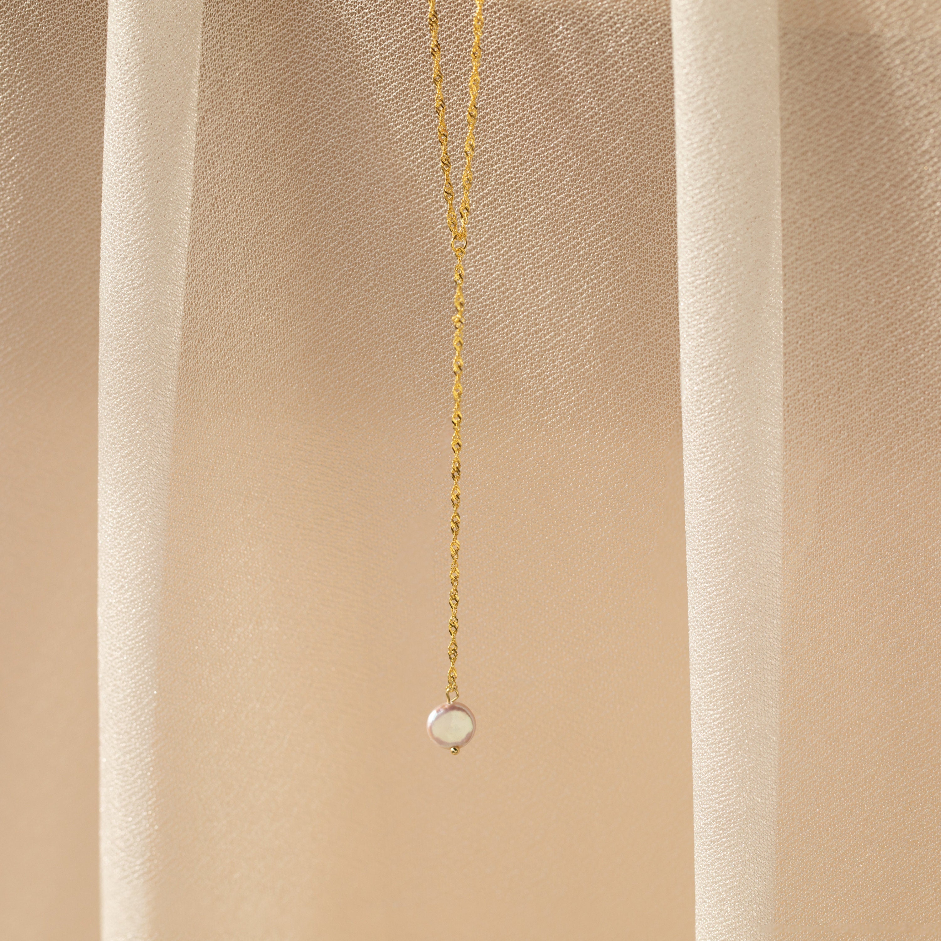 A delicate chain featuring a small round pendant is displayed on a beige, sheer fabric—perfect for fans of timeless elegance like the Pearl Lariat Y Necklace.