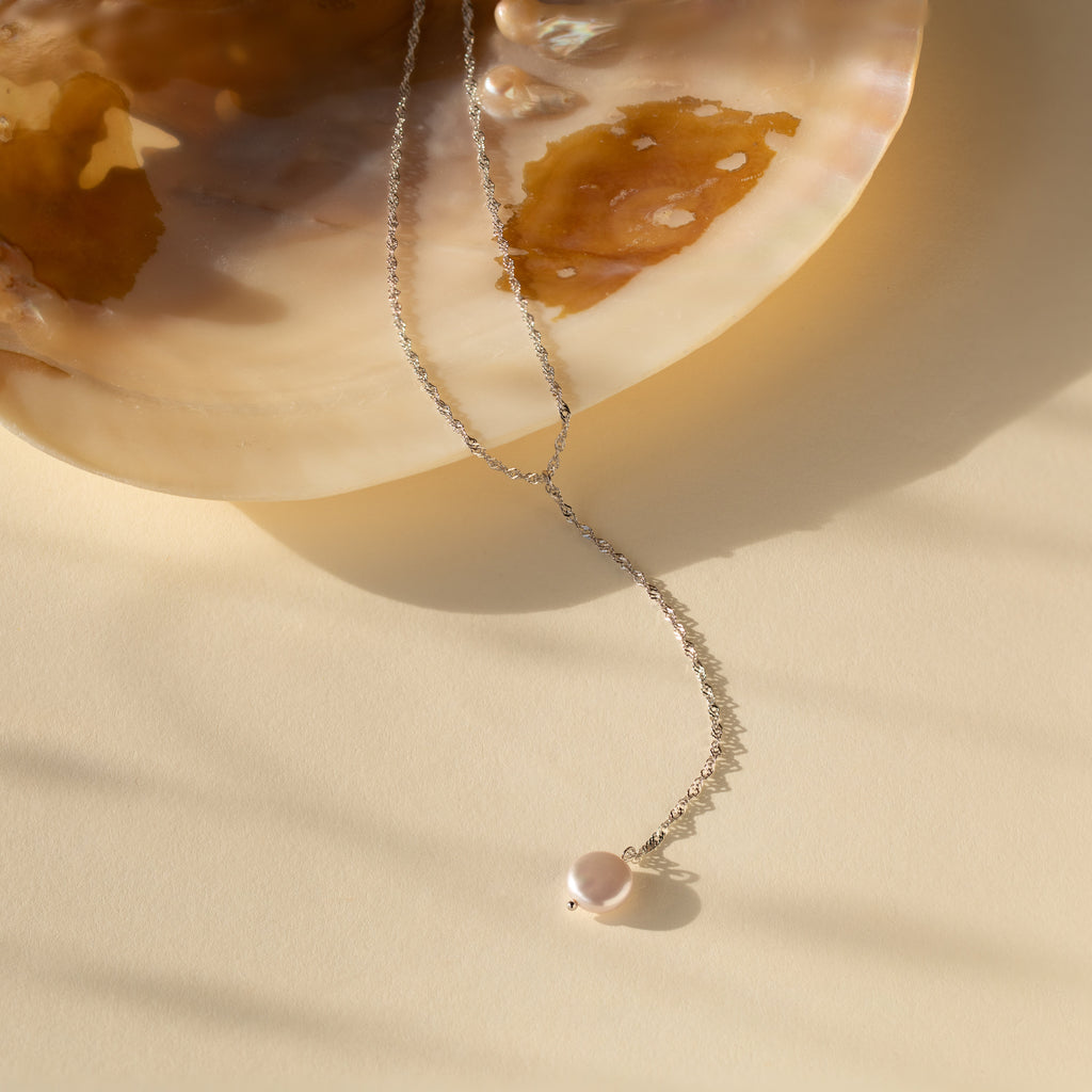 The Pearl Lariat Y Necklace, featuring a delicate chain and a small pearl charm pendant, is displayed on a large seashell against a beige background.