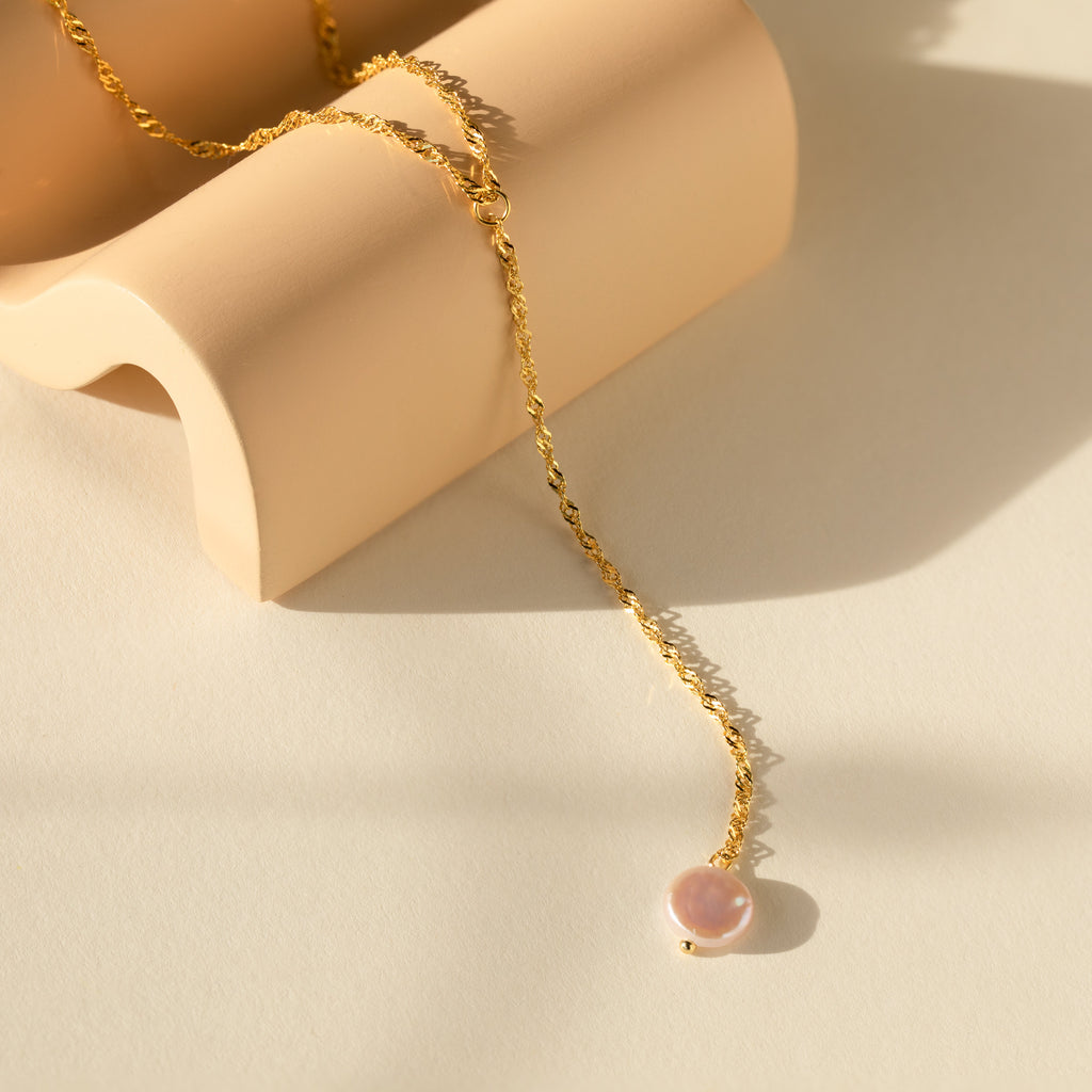 The Pearl Lariat Y Necklace, featuring a delicate chain and round pearl charm, is displayed on a curved beige surface under soft sunlight.