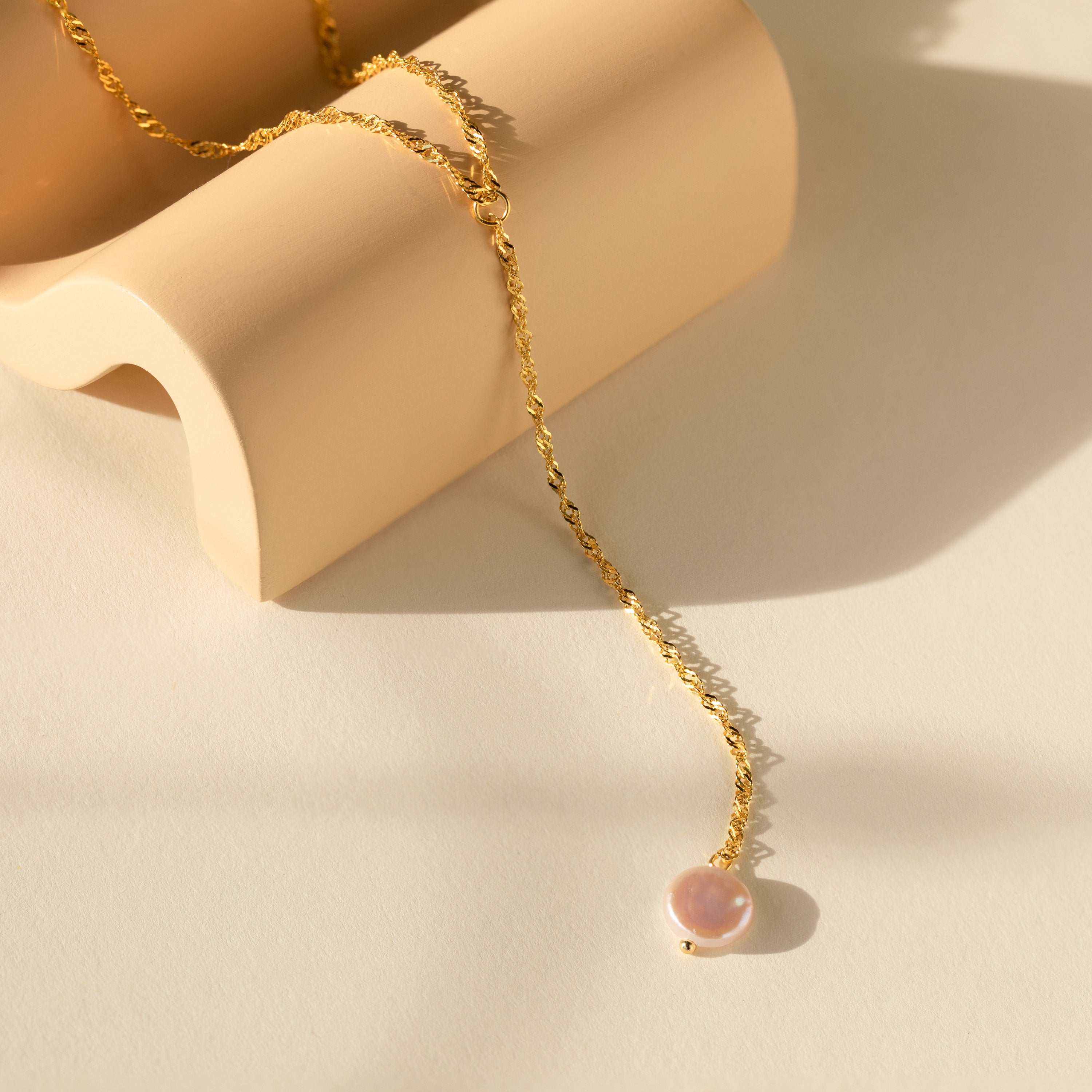 The Pearl Lariat Y Necklace, featuring a delicate chain and round pearl charm, is displayed on a curved beige surface under soft sunlight.