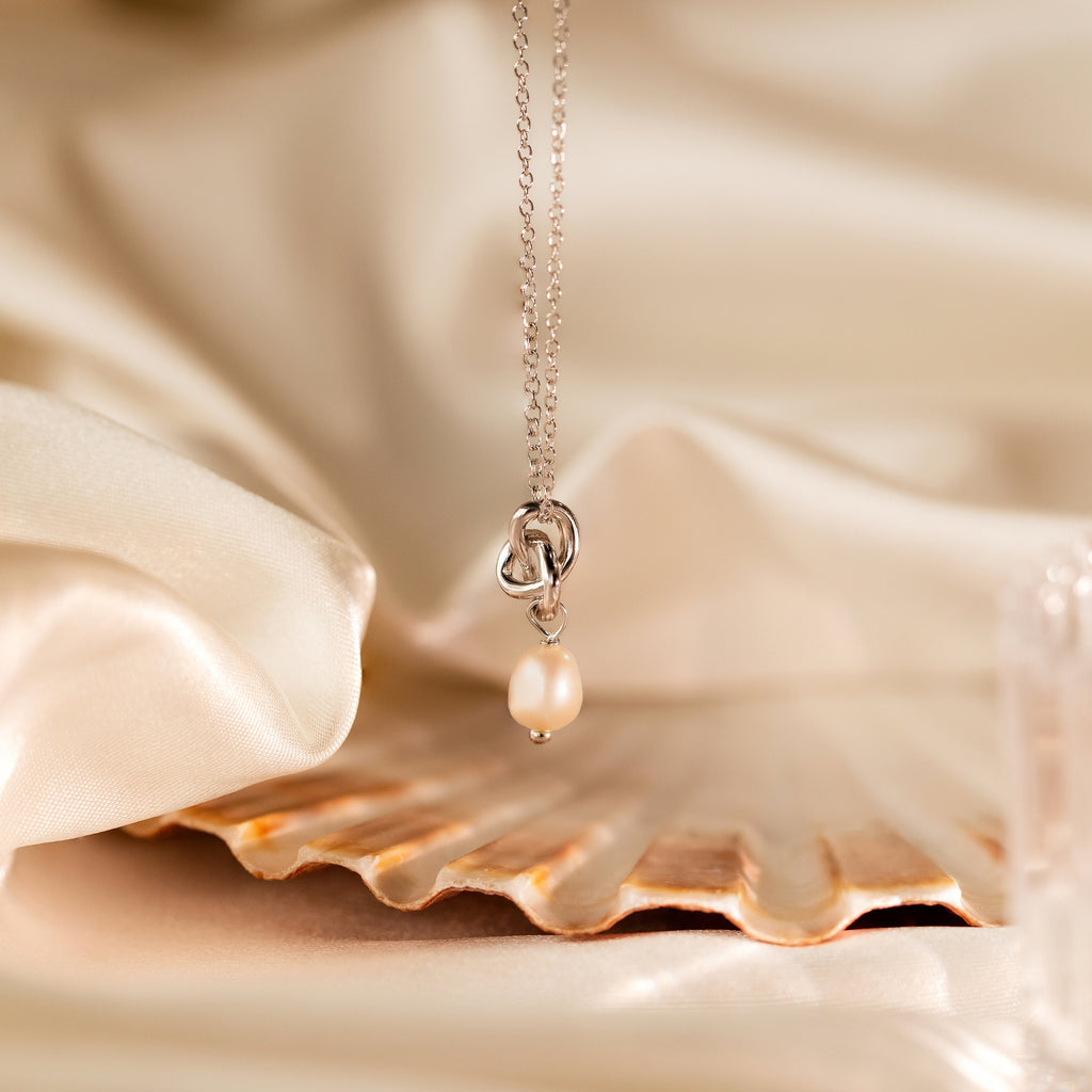The Dainty Knot Pearl Necklace is elegantly showcased on a seashell, highlighting its pearl charm pendant against a soft satin fabric background.