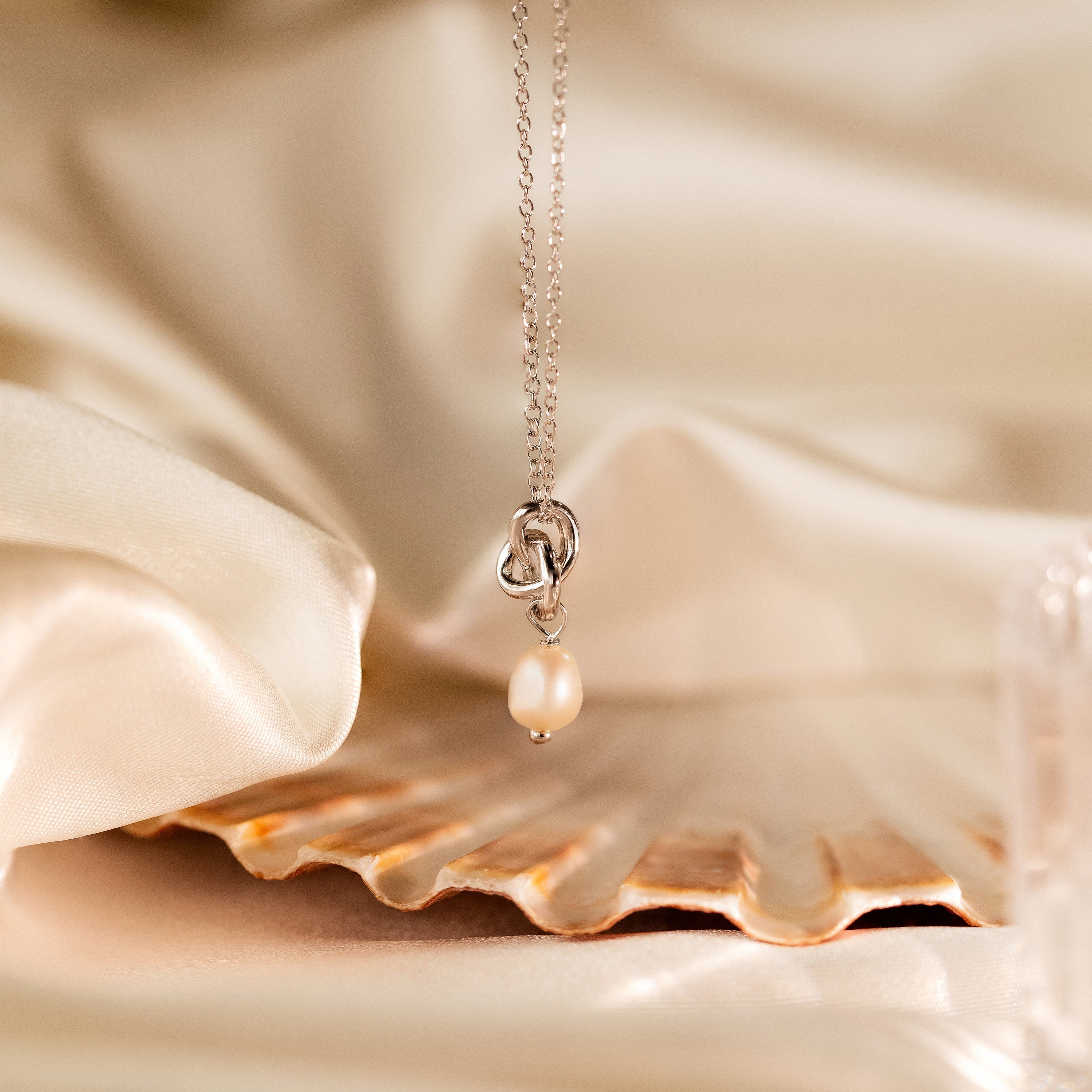 The Dainty Knot Pearl Necklace is elegantly showcased on a seashell, highlighting its pearl charm pendant against a soft satin fabric background.