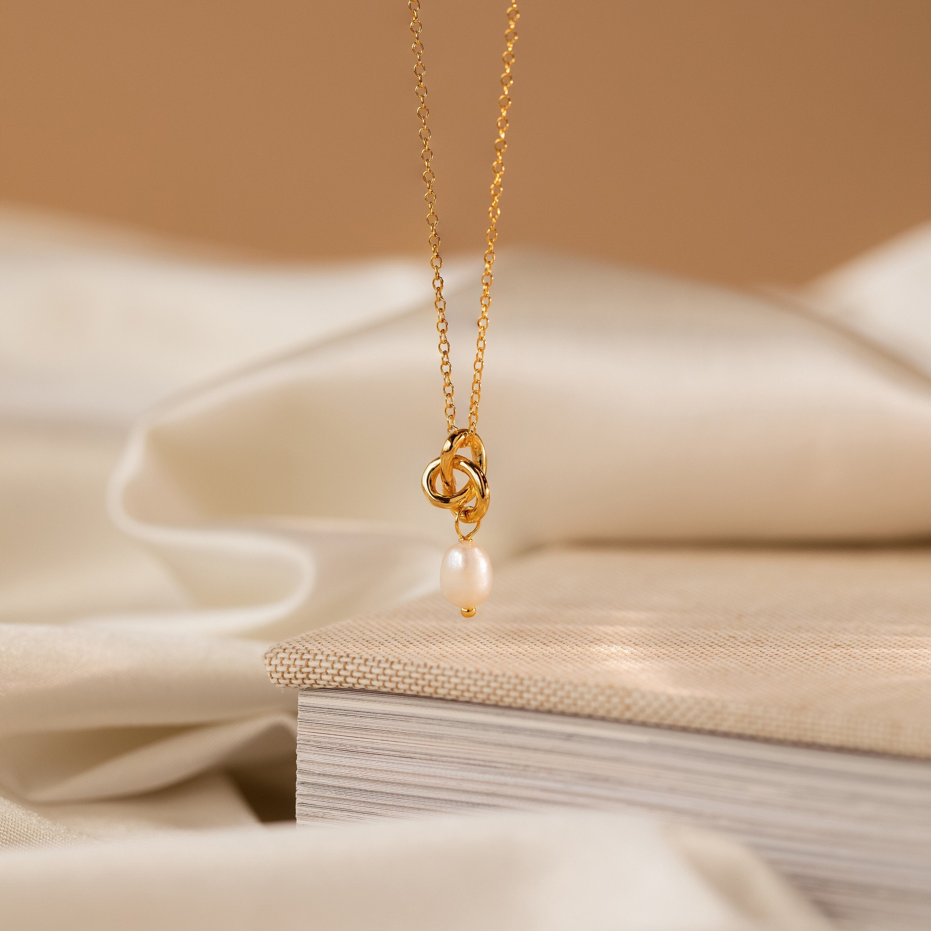 The Dainty Knot Pearl Necklace is draped over a closed book, showcasing its elegant knot design and lustrous pearl charm against a soft fabric background.