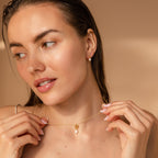 A woman with natural makeup holds the Dainty Knot Pearl Necklace, featuring a pearl charm, and wears matching pearl earrings.