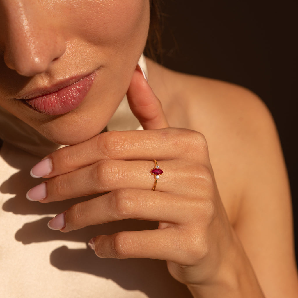 A woman with pink nails showcases the Pave Marquise Birthstone Ring, distinguished by its striking red gemstone, as she holds her hand near her face.