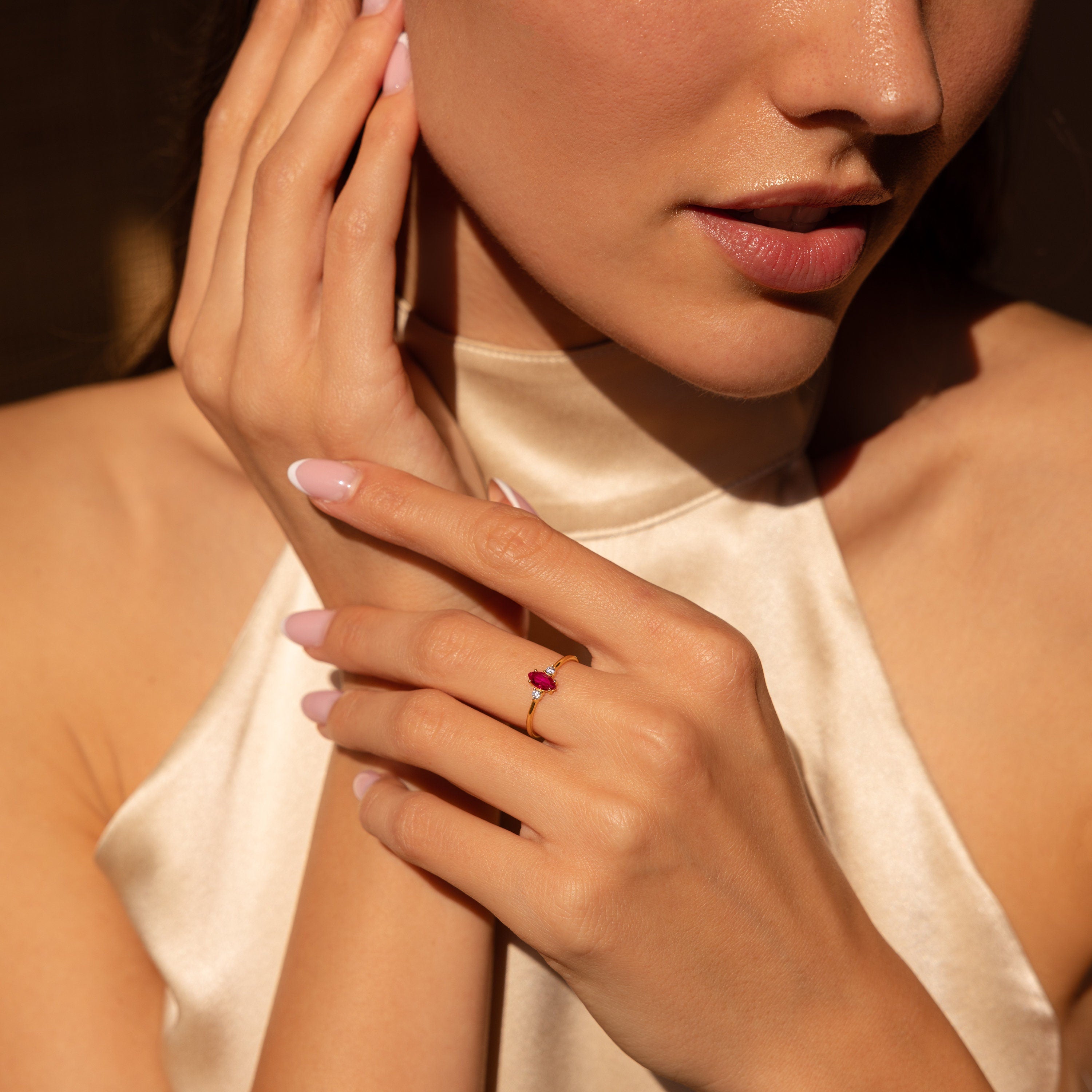 A woman in a satin top touches her face, showcasing the Pave Marquise Birthstone Ring—a gold ring with a red marquise gemstone—adding an elegant touch to her look.