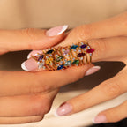 A hand with a French manicure wears stacked gold rings, including a Pave Marquise Birthstone Ring adorned with colorful gemstones, on each finger.