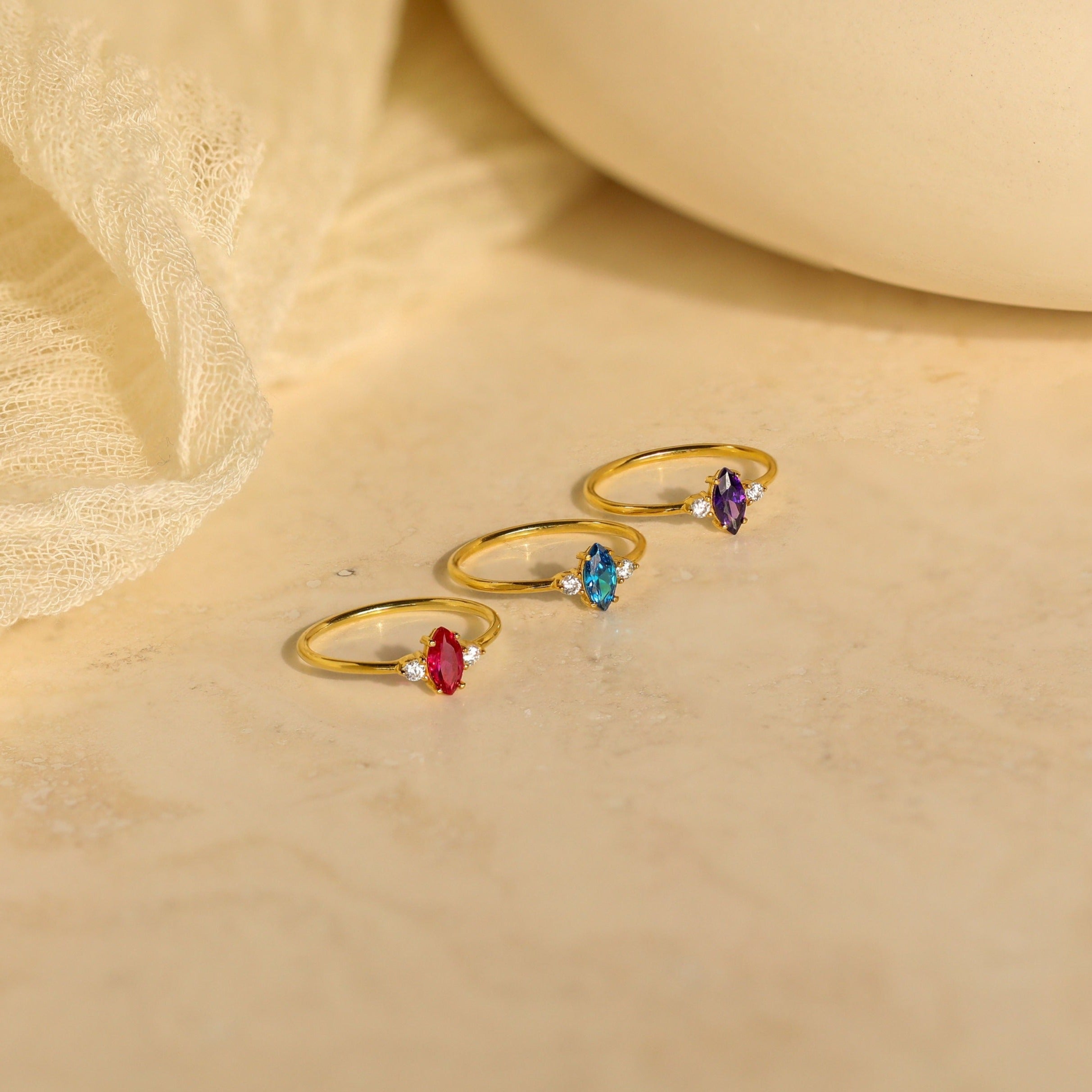 Three Pave Marquise Birthstone Rings with red, blue, and purple personalized stones are displayed on a beige surface with a fabric background—perfect as custom birthstone rings for any special occasion.