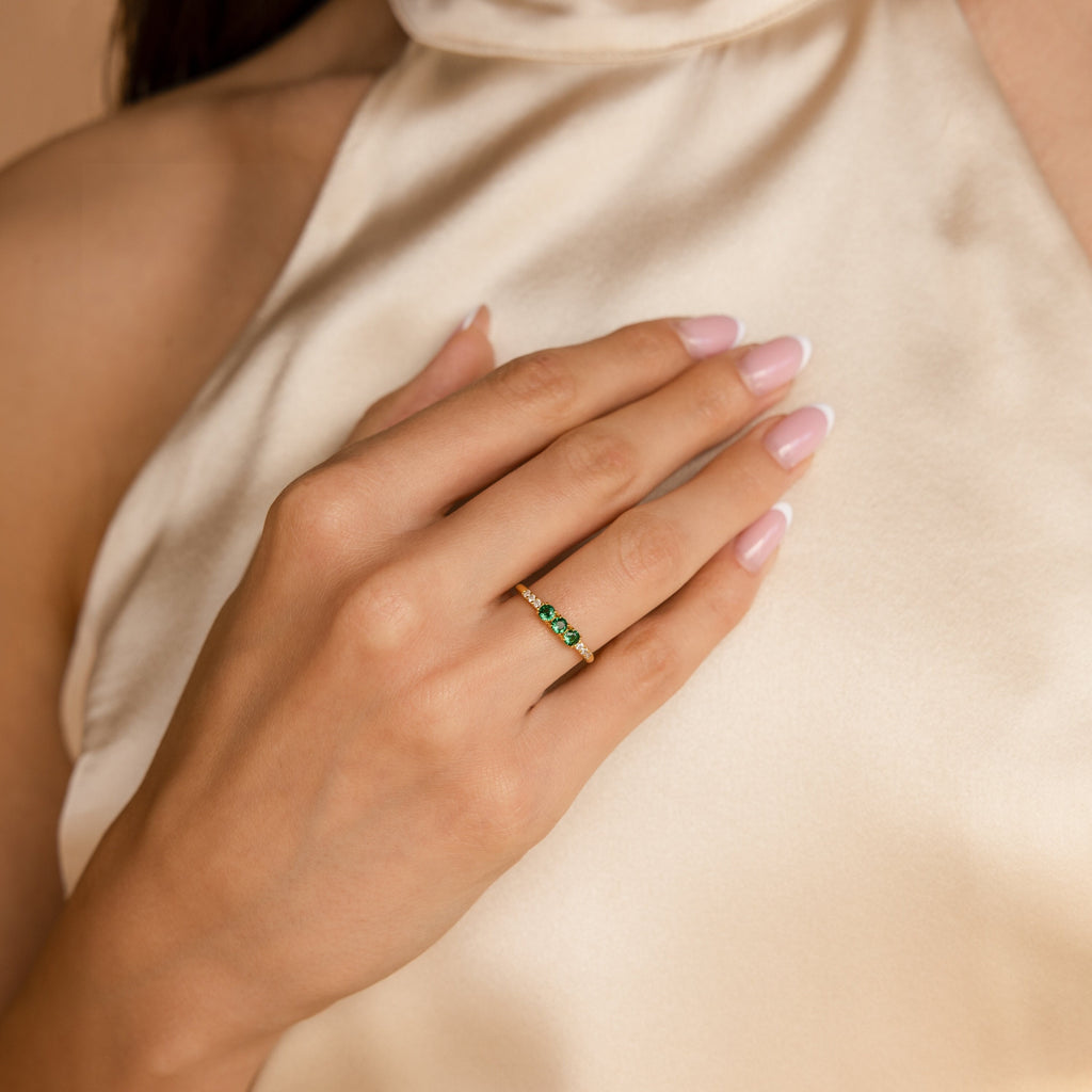 A woman's manicured hand rests on her chest, adorned with the Pave Triple Birthstone Ring—personalized jewelry with green gemstones set in gold.