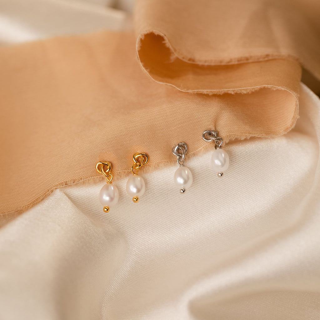 Two pairs of Dainty Knot Pearl Earrings—one gold, one silver—are displayed on cream and peach fabric, exuding classic charm and timeless elegance.