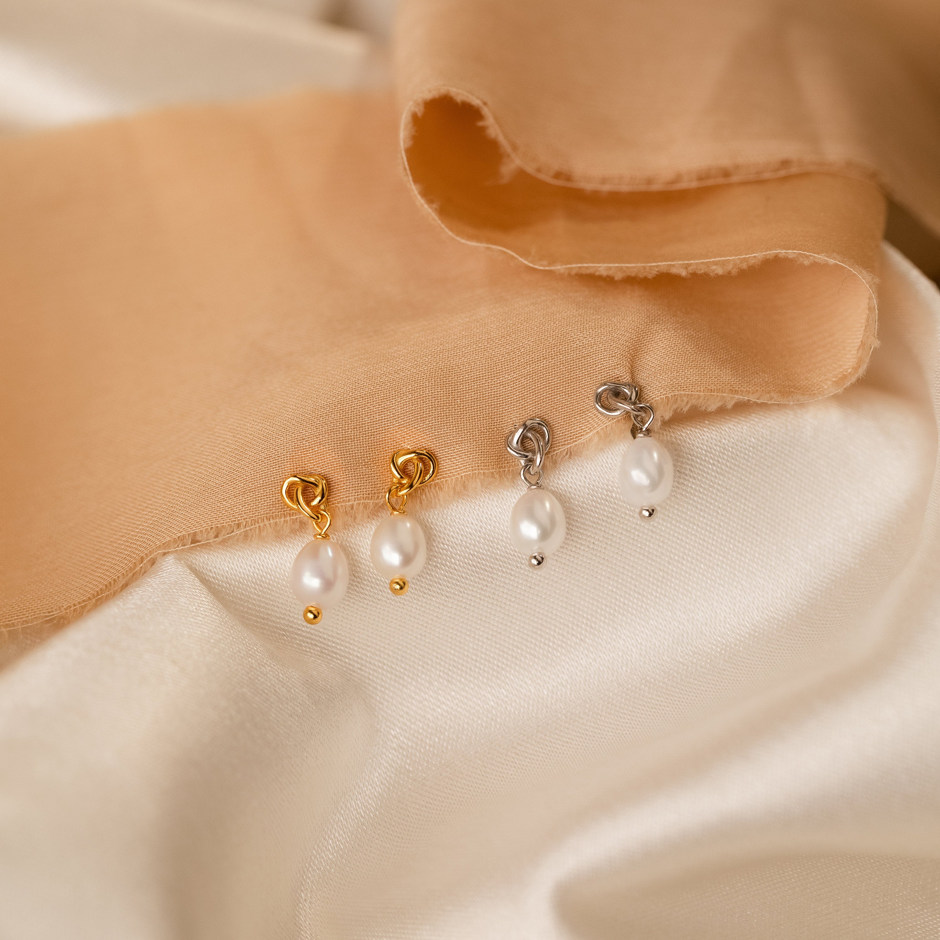 Two pairs of Dainty Knot Pearl Earrings—one gold, one silver—are displayed on cream and peach fabric, exuding classic charm and timeless elegance.