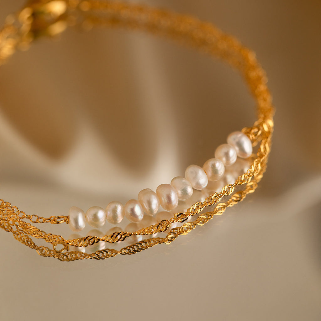 The Duo Pearl Twist Bracelet features a gold minimalist chain with white pearls, showcased on a soft beige background—a chic accessory for effortless elegance.