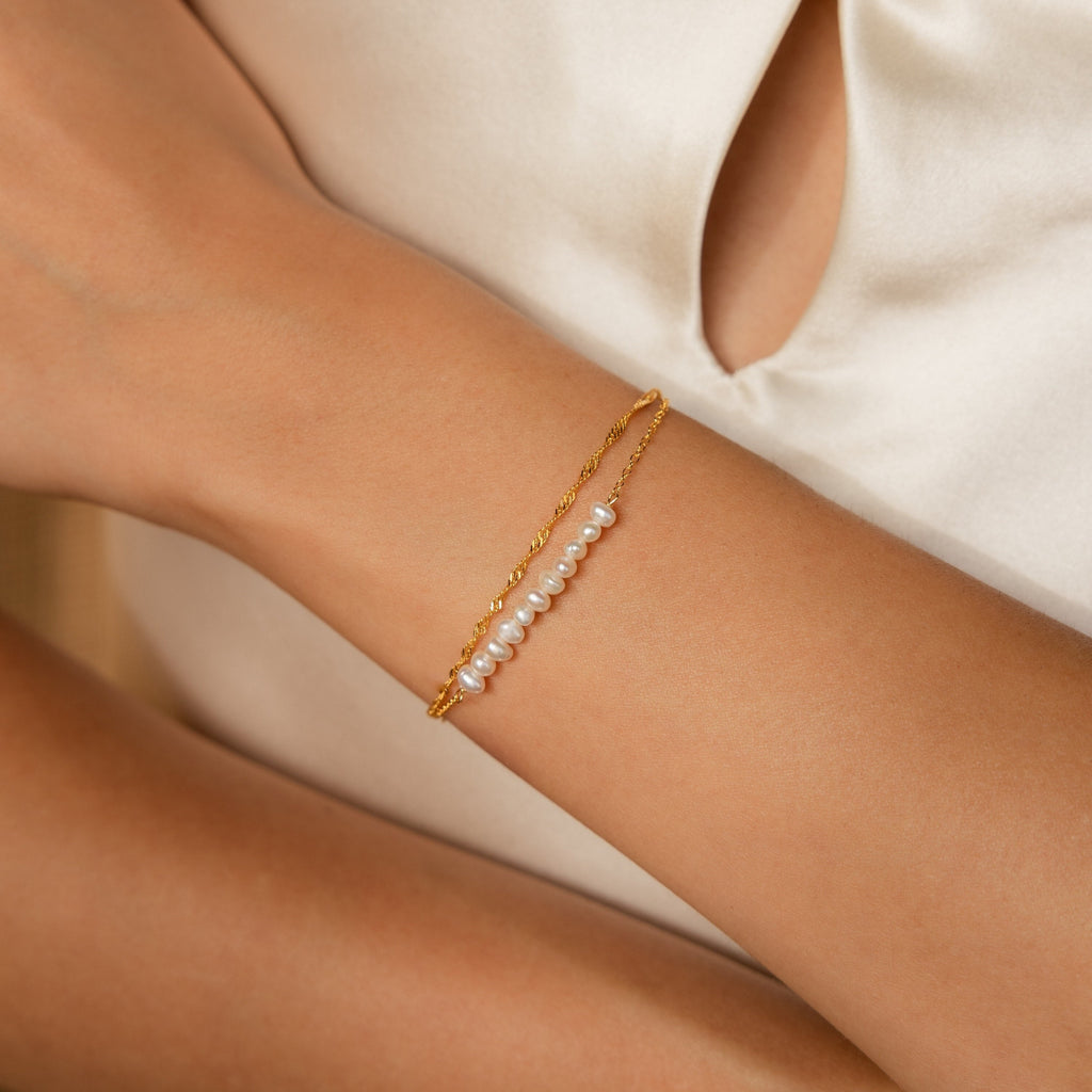 The Duo Pearl Twist Bracelet, a delicate gold minimalist design featuring small pearls, is styled on a woman’s wrist paired with a cream-colored top.