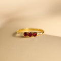 The Pave Triple Birthstone Ring features three red gemstones and small clear stones on the band, shown on a beige surface—ideal for personalized jewelry lovers seeking a customizable birthstone ring.
