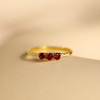 The Pave Triple Birthstone Ring features three red gemstones and small clear stones on the band, shown on a beige surface—ideal for personalized jewelry lovers seeking a customizable birthstone ring.