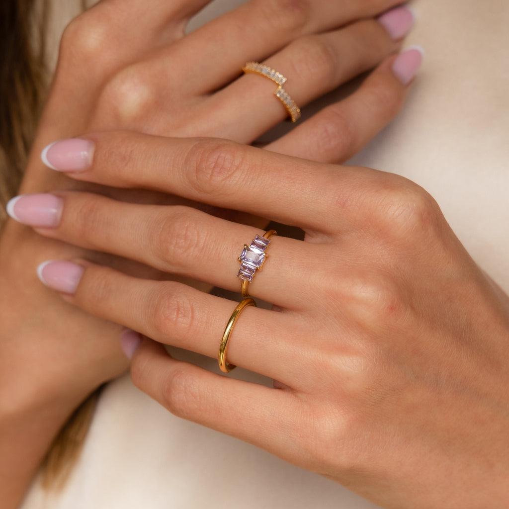 Close-up of hands with light pink nails wearing three gold rings, including the Art Deco Birthstone Ring featuring baguette-cut stones and sparkling diamonds.