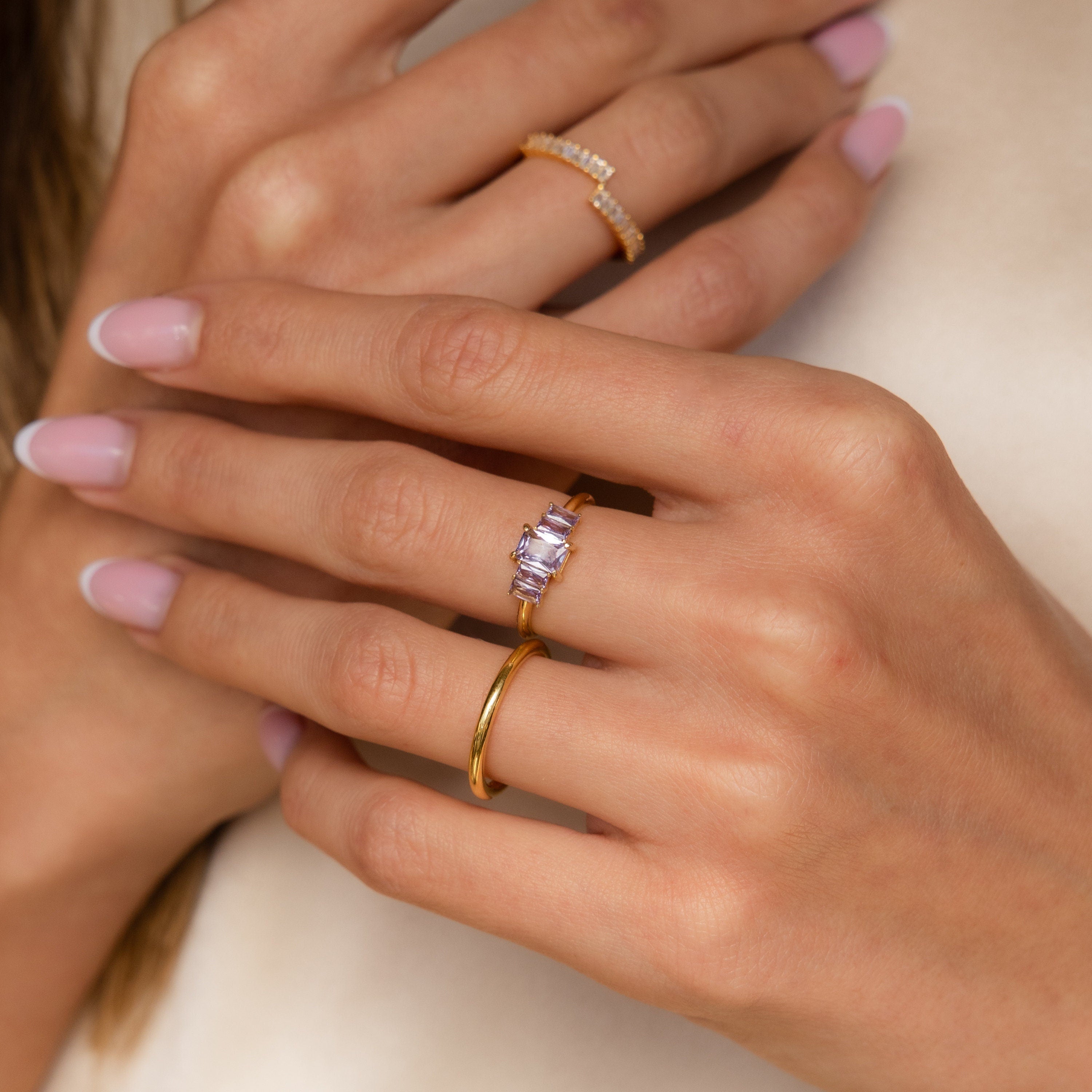Close-up of hands with light pink nails wearing three gold rings, including the Art Deco Birthstone Ring featuring baguette-cut stones and sparkling diamonds.