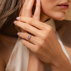 A woman with manicured nails gently touches her face, showcasing the vintage charm of the Art Deco Birthstone Ring adorned with purple baguette-cut stones.