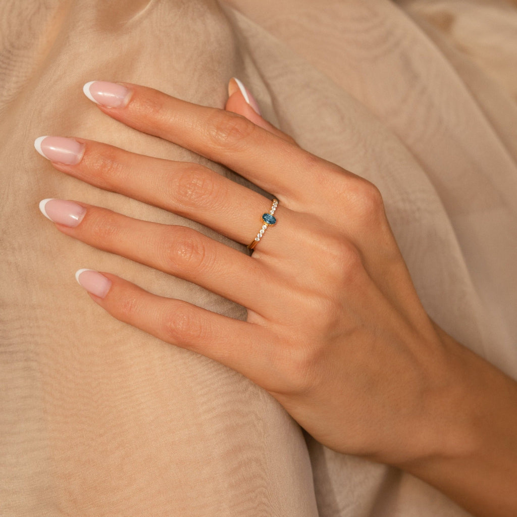 A hand with a French manicure wears the Pave Oval Birthstone Ring, showcasing a blue gemstone and resting elegantly on beige fabric.