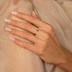 A hand with a French manicure wears the Pave Oval Birthstone Ring, showcasing a blue gemstone and resting elegantly on beige fabric.