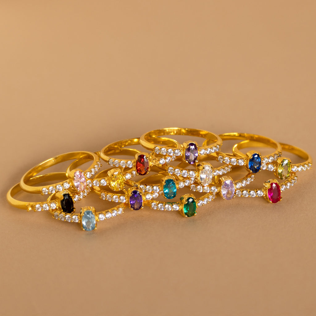A Pave Oval Birthstone Ring, featuring a gold band with a colorful gemstone center and small clear stones, is displayed on a tan surface.