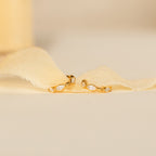 The Marquise Inlay Huggies—gold hoops with small, bezel-set gemstones—rest elegantly on light, sheer fabric.